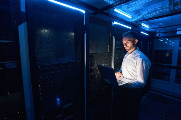 Hombre caucásico en el centro de datos de pie cerca de los bastidores de servidores con equipos de red y usando una computadora portátil