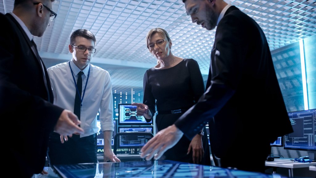 Team of Corporate Managers Working at the Table in Monitoring Room.  They're in State of the Art Facility. Computers have Animated Screens.