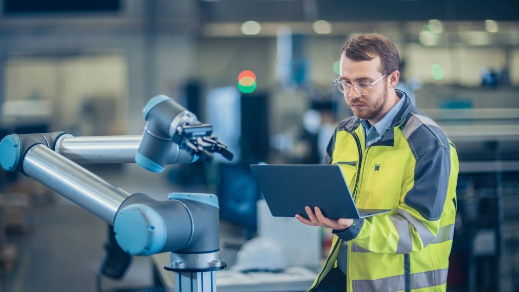 At the Factory: Automation Engineer Uses Laptop for Programming Robotic Arm. New Era in Automatic Manufacturing Industry.